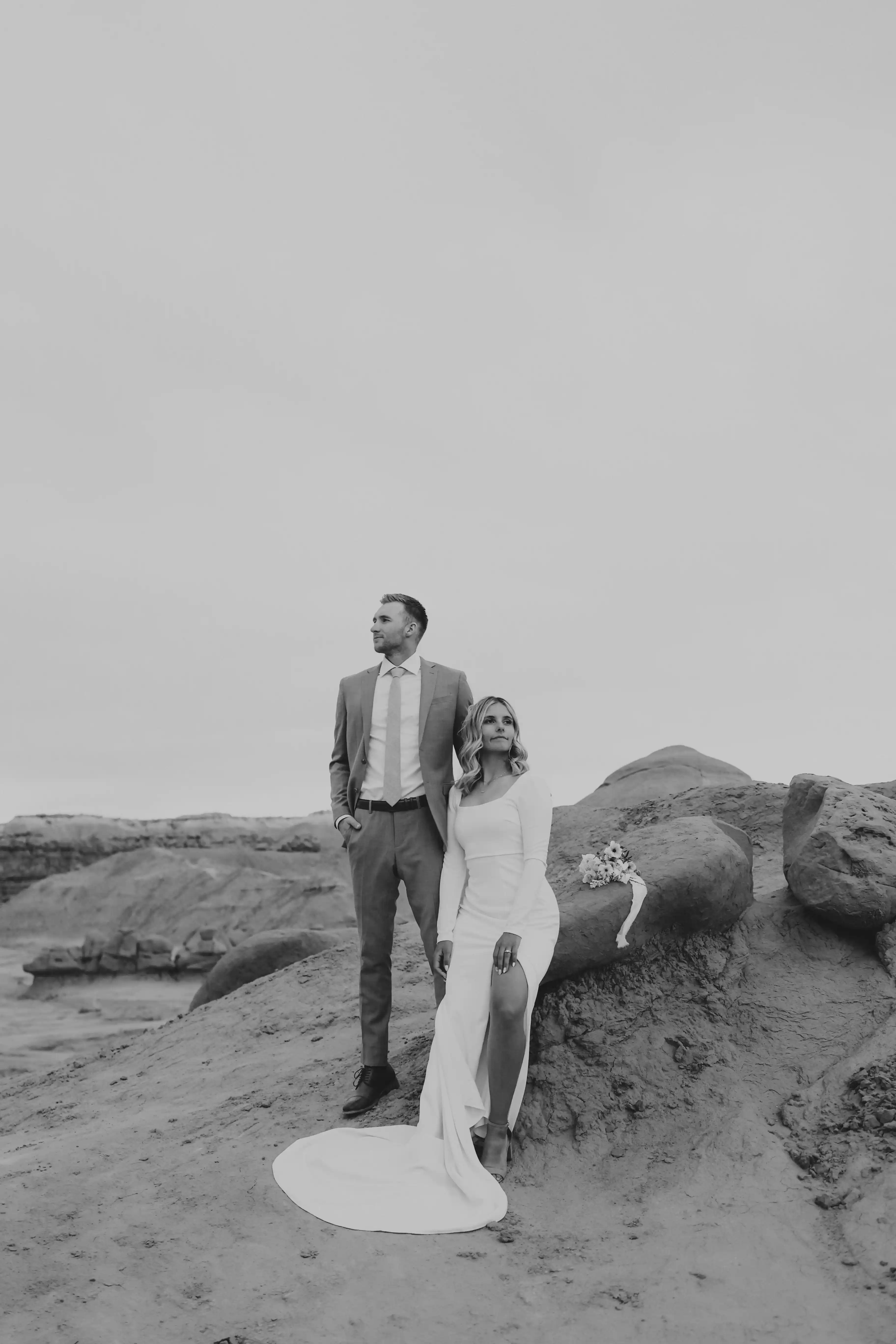 Bride sitting on rock with groom standing and looking in the distance